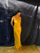 A woman in the Mila Halter Silk Dress - Marigold by Rimmba | Sustainable Clothing stands barefoot on a woven mat, gazing down, with a dark textured backdrop and dappled sunlight.