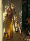 A woman wears the Rimmba Mila Halter Silk Dress in Marigold, standing barefoot on a woven mat by a sunlit wall with leafy shadows, surrounded by lush outdoor greenery.