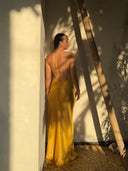 A woman in the Mila Halter Silk Dress - Marigold by Rimmba | Sustainable Clothing stands in a sunlit corner near a wooden ladder, with leaf shadows on the wall and woven flooring beneath her feet.