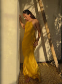 A woman in the Mila Halter Silk Dress - Marigold by Rimmba | Sustainable Clothing stands barefoot on a woven mat, gently holding her dress as sunlight and leafy shadows create a serene, summery mood.