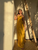 A woman, barefoot and bathed in sunlight and soft shadows, stands by a wooden ladder in the Mila Halter Silk Dress - Marigold from Rimmba | Sustainable Clothing, leafy patterns dancing across the white wall beside her.