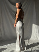 A woman models the Rimmba Mila Halter Silk Dress in White Ivory, featuring a backless design, halter neckline, and side slit, standing sideways in sunlight against a draped white fabric background.