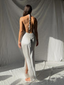 A woman stands barefoot on a white fabric backdrop, wearing the Mila Halter Silk Dress - White Ivory Bridal Dress by Rimmba | Sustainable Clothing. Sunlight casts distinct shadows across the draped background.