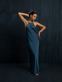 A woman models the Mila Halter Silk Dress - Indigo by Rimmba | Sustainable Clothing, posing barefoot against a dark backdrop with one arm bent over her head and wearing a floral headpiece.
