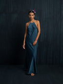 A woman stands barefoot against a dark textured background, wearing the Rimmba Mila Halter Silk Dress in Indigo. Her hair is slicked back and adorned with a small purple flower.
