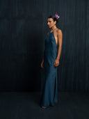 A woman poses against a dark background in the Rimmba Mila Halter Silk Dress - Indigo, featuring an open back and side slit. She has a flower arrangement in her hair and stands with her arms relaxed by her sides.