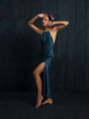 A barefoot woman poses gracefully against a dark background, wearing the Rimmba Mila Halter Silk Dress in Indigo with a thigh-high slit and flowers behind her ear.