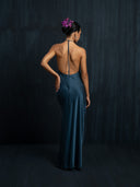 A woman with her hair in a bun adorned with purple flowers faces away, wearing the Rimmba Mila Halter Silk Dress in Indigo, set against a dark, textured background.
