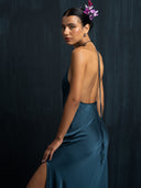 A woman in the Rimmba Mila Halter Silk Dress in indigo stands against a dark background, looking over her shoulder with a pink flower in her hair and a calm, confident expression.