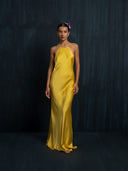 A woman poses before a dark, textured backdrop wearing the Mila Halter Silk Dress in marigold by Rimmba | Sustainable Clothing. Her hair is slicked back with a pink flower accent, and she has a neutral expression.