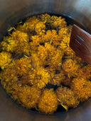 Bright orange marigold flowers, echoing the Marigold hue of Rimmba's Mila Halter Silk Dress, float in water within a metal pot, with a wooden utensil partially seen on the right.