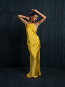 A woman poses gracefully barefoot against a dark textured background, wearing the Rimmba | Sustainable Clothing "Mila Halter Silk Dress - Marigold," with flowers in her hair and a serene expression.