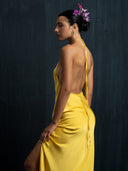 A woman models the Mila Halter Silk Dress - Marigold by Rimmba | Sustainable Clothing, posing sideways against a dark background with an elegant updo adorned with purple flowers and a serene gaze.