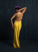 A woman, back to the camera, wears the Mila Halter Silk Dress in marigold by Rimmba | Sustainable Clothing and a purple floral headpiece. She raises an arm to touch her dress strap against a dark, plain background.