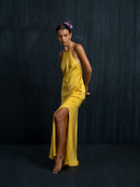 A woman poses barefoot against a dark textured background, wearing the Mila Halter Silk Dress in marigold from Rimmba | Sustainable Clothing, with purple flowers adorning her hair.