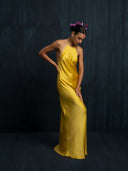 A woman in the Mila Halter Silk Dress - Marigold by Rimmba | Sustainable Clothing stands against a dark background, tilting her head down, touching her hip, with purple flowers in her hair and an elegant posture.