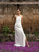 Barefoot woman standing in front of natural wall wearing Mulberry silk halter maxi dress white. Hancrafted botanical-dyed maxi dress sustainable luxury fashion for bridal and resort wear.