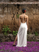 Back-view of woman is facing a natural wall wearing Mulberry silk Halter dress with backless style and slit on the left side. Hancrafted botanical-dyed maxi dress sustainable luxury fashion for bridal and resort wear.