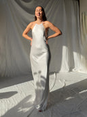 A woman poses in bright sunlight with eyes closed and hands on hips, wearing the Rimmba Mila Halter Silk Dress – a long, white ivory bridal gown – against a white draped fabric background.