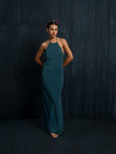 A woman stands barefoot against a dark textured background, gazing down with hands clasped. She wears the Mila Halter Silk Dress in emerald green crepe by Rimmba | Sustainable Clothing and has flowers in her hair.