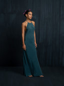 A woman stands barefoot against a dark textured background in the Rimmba Mila Halter Silk Dress—Emerald Green Crepe. Her dark hair is styled up with pink flowers, and she gazes ahead with a calm expression. Sustainable clothing by Rimmba.