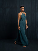 A woman stands barefoot against a dark backdrop, wearing the Mila Halter Silk Dress in emerald green crepe by Rimmba | Sustainable Clothing, and a headpiece adorned with purple flowers. She gazes forward with a neutral expression.