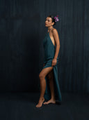 A woman in the Mila Halter Silk Dress - Emerald Green Crepe by Rimmba | Sustainable Clothing stands barefoot against a dark backdrop, a pink flower in her hair, posing gracefully with eyes closed to create an elegant, serene mood.