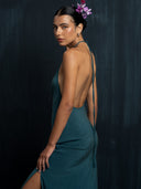 A woman in the Mila Halter Silk Dress - Emerald Green Crepe by Rimmba | Sustainable Clothing stands sideways against a dark background, looking over her shoulder, with dark hair styled neatly and a pink flower behind her ear.