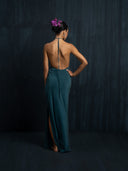 A woman, barefoot and with her hair in a bun adorned with purple flowers, stands against a dark textured background wearing the Rimmba Mila Halter Silk Dress in emerald green crepe.