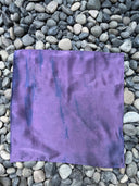 The Mini Silk Scarf by Rimmba | Sustainable Clothing, a square purple silk satin piece with subtle blue streaks and natural dyes, is displayed flat on a surface of gray and black smooth stones.