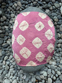 A large smooth gray rock is draped with the Mini Silk Scarf by Rimmba | Sustainable Clothing, featuring pink silk satin with white geometric patterns, surrounded by smaller gray pebbles.