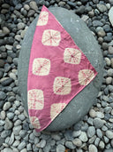 A large smooth gray oval rock rests among pebbles, topped with the Rimmba | Sustainable Clothing Mini Silk Scarf in pink with a tan geometric pattern.