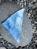 The Mini Silk Scarf by Rimmba | Sustainable Clothing, in blue tie-dye silk satin and folded into a triangle, is displayed on a large gray stone amid small rounded pebbles.