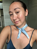 A woman with tied-back hair wears a blue top and the Mini Silk Scarf from Rimmba | Sustainable Clothing around her neck. She smiles slightly at the camera indoors, against a neutral background with grey walls.