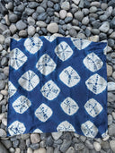 The Rimmba Mini Silk Scarf, handcrafted with natural dyes, features blue and white geometric tie-dye patterns and diamond shapes. Shown laid flat on smooth gray stones. Sustainable fashion by Rimmba | Sustainable Clothing.