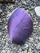 A large, smooth rock on smaller stones is draped with the Mini Silk Scarf by Rimmba | Sustainable Clothing—a handcrafted silk satin scarf dyed purple with darker diagonal streaks using natural dyes.