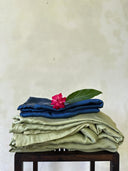 The Mint Green - Full Silk Bedding Set by Rimmba | Sustainable Clothing is artfully folded atop a dark wooden table, adorned with a green leaf and bright pink flower, set against a plain off-white wall.