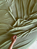 A hand gently grasps the soft, light green Mulberry Silk of the Mint Green - Full Silk Bedding Set by Rimmba | Sustainable Clothing, creating elegant folds—an exquisite touch to any botanical silk bedding.