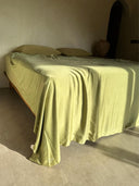 A bed dressed in Rimmba's Mint Green - Full Silk Bedding Set with two pillows sits in a sunlit room with minimalist beige walls, soft shadows on the floor, and a large vase and dark doorway in the background.