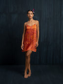 A woman stands barefoot against a dark textured background, wearing the Mira Eco-Print Dress - Mini by Rimmba | Sustainable Clothing. Her hair is pulled back with a small pink flower tucked behind her ear.
