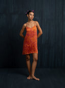 A woman stands barefoot against a dark textured backdrop, wearing the Mira Eco-Print Dress - Mini by Rimmba | Sustainable Clothing. She has flowers in her hair and her arms are relaxed behind her back.
