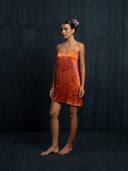 A woman stands barefoot against a dark textured background, wearing the Mira Eco-Print Dress - Mini by Rimmba | Sustainable Clothing, featuring sleeveless hand-dyed orange and red patterns. Her hair is pulled back with a purple flower accessory.