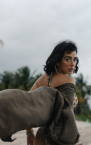 A stylish woman is standing on a sandy beach and looking back over her shoulder. She is wearing a patterned swimsuit with a Wabisabi kimono mud cover-up that is blowing dramatically in the wind. Her short dark hair frames her face, and she is wearing large hoop earrings and bracelets.