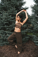 A woman is posing gracefully outdoors among tall evergreen trees. She is wearing Pevita halter top mud and akasha  pants, creating a natural and earthy look. Her arms are raised above her head in a dance-like pose, while one leg is slightly lifted, giving the image an artistic and elegant feeling.