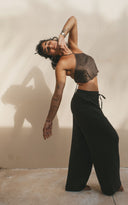 Hannah Singer's  is posing artistically against a plain light-colored wall, creating a soft and minimalist atmosphere. She is wearing Pevita halter mud top and Akasha pants. Her body is bent gracefully to the side, with one arm curved above her head and the other hanging naturally, giving the pose a fluid, dance-like elegance.
