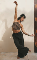 Hannah Singer's  posing gracefully in a minimalist indoor setting, appearing to perform a contemporary dance movement. She is standing barefoot against a light-colored wall, with one arm stretched upward and the other extended downward in a fluid, elegant gesture. She wears Pevita silk mud top and akasha mud pants.