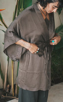 A woman is standing indoors while adjusting the belt of her loose Mud Kimono. She is wearing dark pants and several accessories, including large hoop earrings, bracelets, and turquoise rings. Her short curly hair frames her face as she looks down with a focused expression.