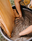 The image shows two people using their hands to mix thick mud inside a large black container. One person is wearing a patterned garment, while pieces of brown fabric are soaked in the muddy mixture nearby. The scene appears to capture a hands-on, traditional fabric dying making process.