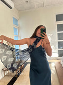A woman takes a mirror selfie in a sunlit room, wearing a dark dress and wrapping the Night Shibori - Indigo Silk Chiffon Scarf by Rimmba | Sustainable Clothing around her neck. The hand-dyed scarf flows as she stands near a window, phone in hand.