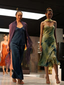 Three models walk a runway—one in Rimmba’s Nindy Silk Pant in Indigo, another in a green tie-dye look, and the third in vibrant orange. A sheer purple shawl adds sophisticated flair to the stylish indoor scene.